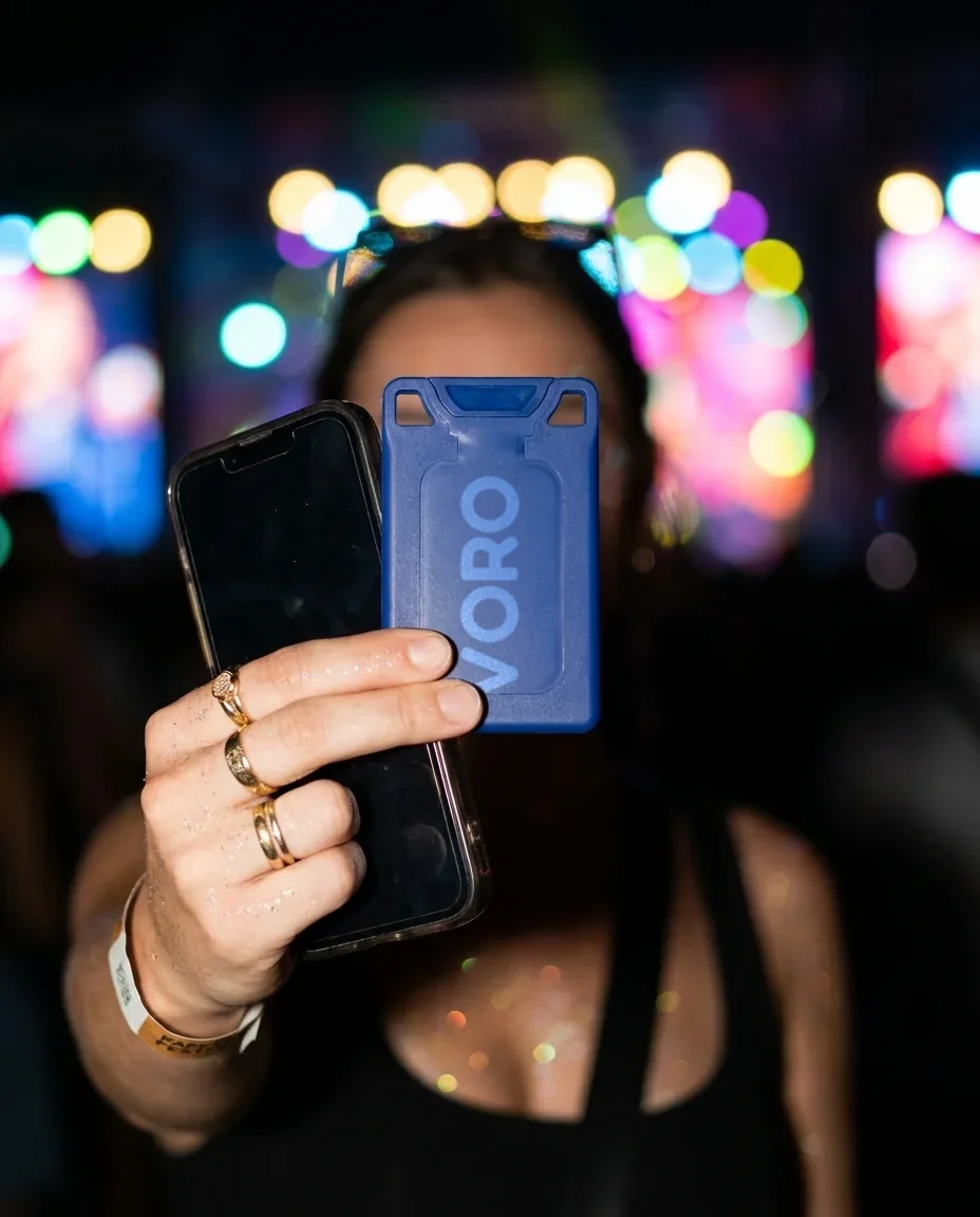 A hand with a festival wristband holding the navy VORO card next to a phone at a concert.